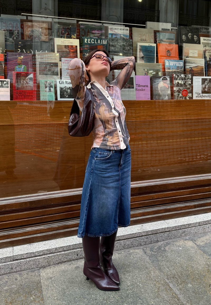 Žena v okuliaroch, oblečená v blúzke s potlačou, rifľovej sukni a hnedých topánkach, stojí pred veľkým oknom, za ktorým sú rôzne knihy a časopisy.