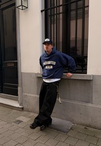 Jeune homme en sweat à capuche bleu marine et casquette de baseball appuyé contre le rebord d'une fenêtre sur un trottoir urbain, avec en arrière-plan une pierre grise et une porte noire.
