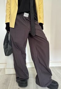 Personne portant un blazer jaune, une chemise noire avec une cravate, un pantalon ample rayé noir et gris, des sabots noirs, et tenant un sac noir à pois blancs.