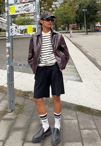 Een persoon draagt een glanzende bordeauxrode jas over een gestreept shirt, zwarte shorts en stevige zwarte loafers. Witte sokken hebben zwarte strepen.