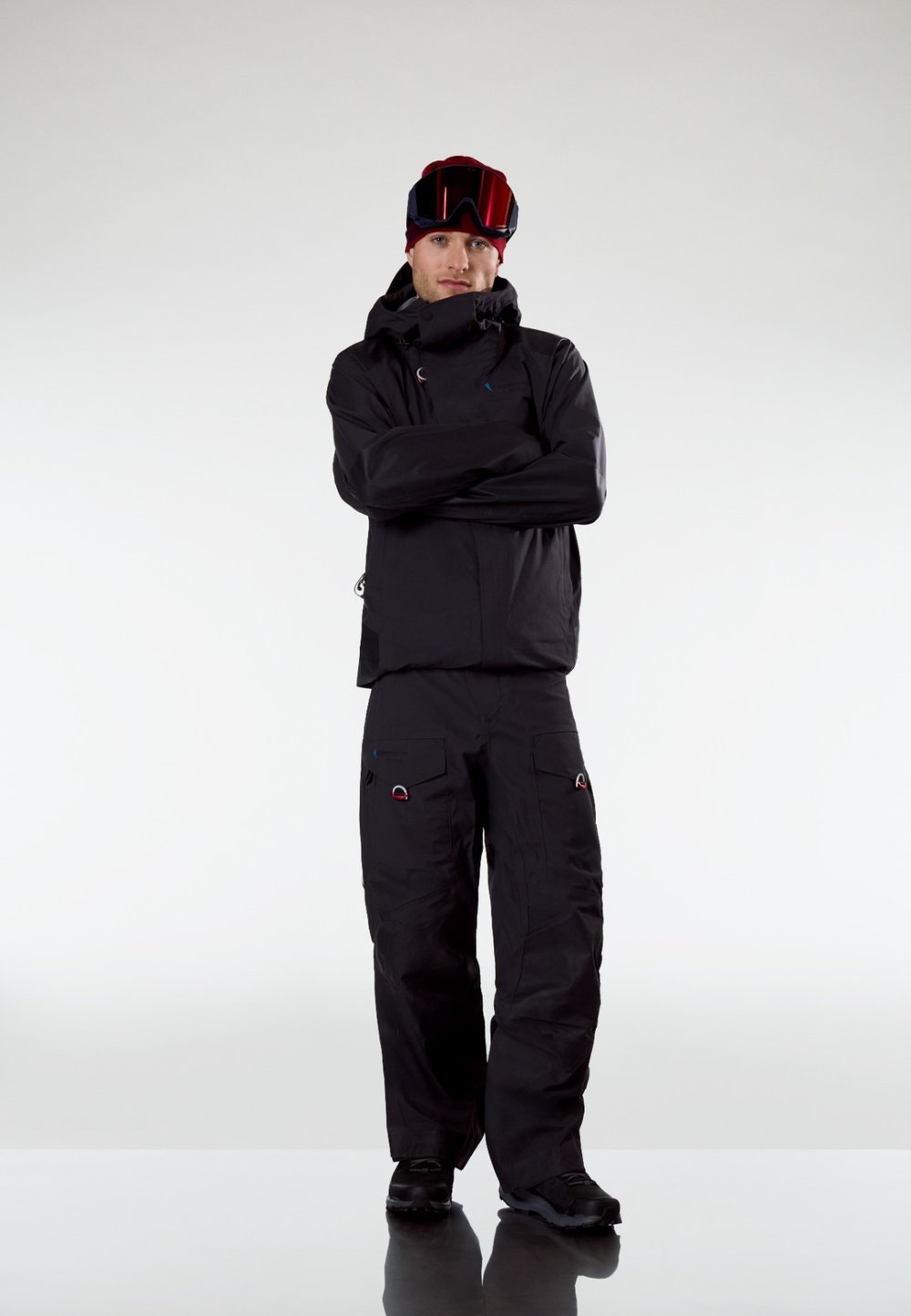 Άνδρας με μαύρο χειμερινό μπουφάν και παντελόνι, κόκκινο σκουφάκι και γυαλιά σκι στέκεται με στα υρωμένα χέρια μπροστά σε ανοιχτόχρωμο απλό φόντο.