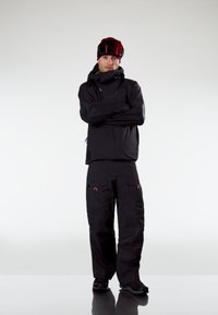 Άνδρας με μαύρο χειμερινό μπουφάν και παντελόνι, κόκκινο σκουφάκι και γυαλιά σκι στέκεται με σταυρωμένα χέρια μπροστά σε ανοιχτόχρωμο απλό φόντο.