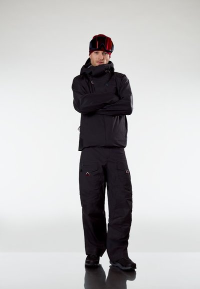 Hombre con chaqueta y pantalones de invierno negros, gorro rojo y gafas de esquí, de pie con los brazos cruzados frente a un fondo claro y liso.