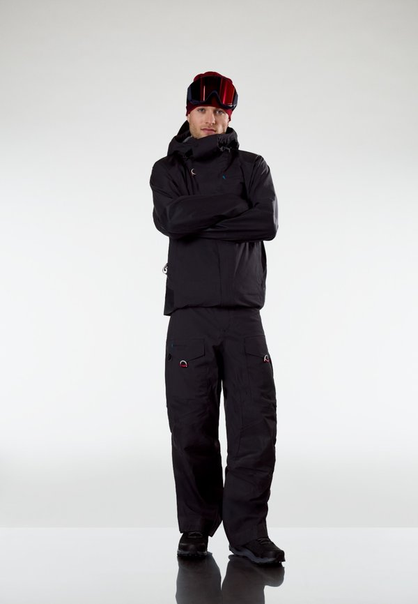 Mees, kes kannab musta talvejope ja pükse, punast mütsi ning suusaprille, seisab käed rüpes lihtsa heleda tausta ees.