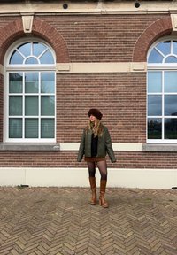 Olijfgroene oversized jas, zwarte trui, bruine minirok en bruin kniehoge laarzen gedragen tegen een bakstenen muur met ramen op de achtergrond.