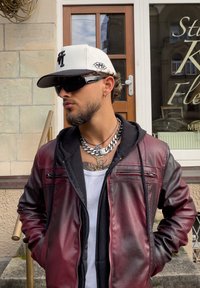 Veste en cuir bordeaux avec accents noirs, T-shirt blanc, colliers en chaîne argentés, casquette blanche et noire, lunettes de soleil foncées.