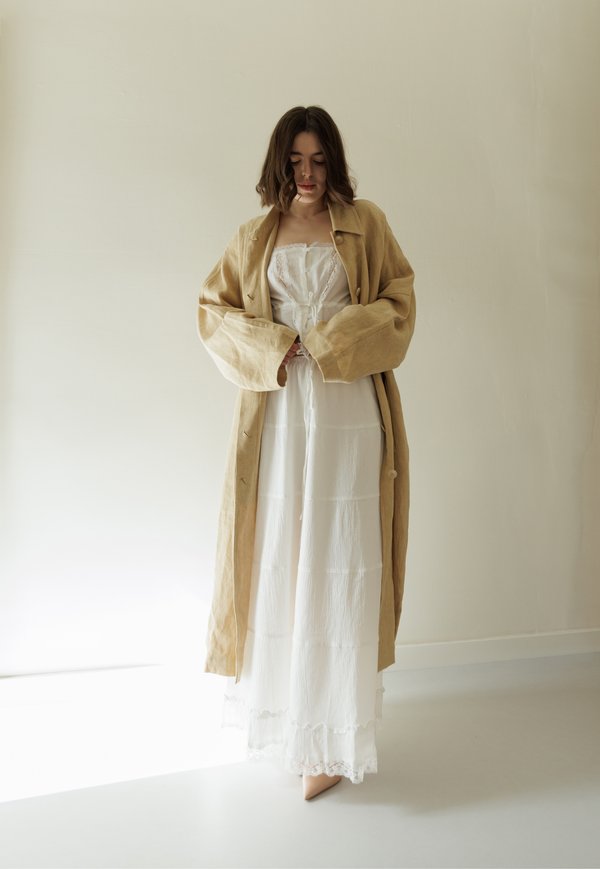 Palton din in bej cu mâneci late peste o rochie maxi albă cu dantelă, care are un corset legat. Texturi fine, culori naturale.
