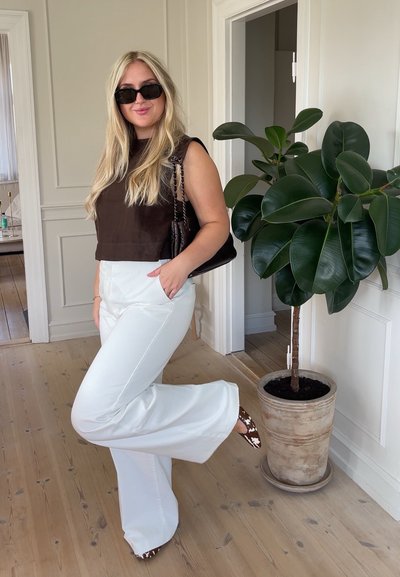 Top corto marrón, pantalones acampanados blancos y tacones altos con estampado. Gafas de sol negras, bolso de hombro y planta verde de interior en el fondo.