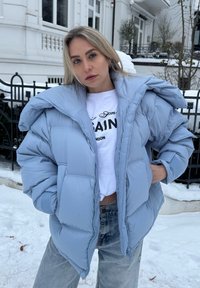 Femme blonde en plein air dans la neige, portant une doudoune bleu clair, un crop top blanc avec un texte, et un jean bleu clair, debout les mains dans les poches.
