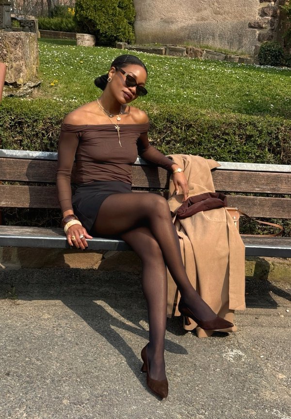 Femme en lunettes de soleil portant un haut décolleté, une jupe noire et des talons, assise sur un banc de parc avec un manteau marron clair à côté d'elle.