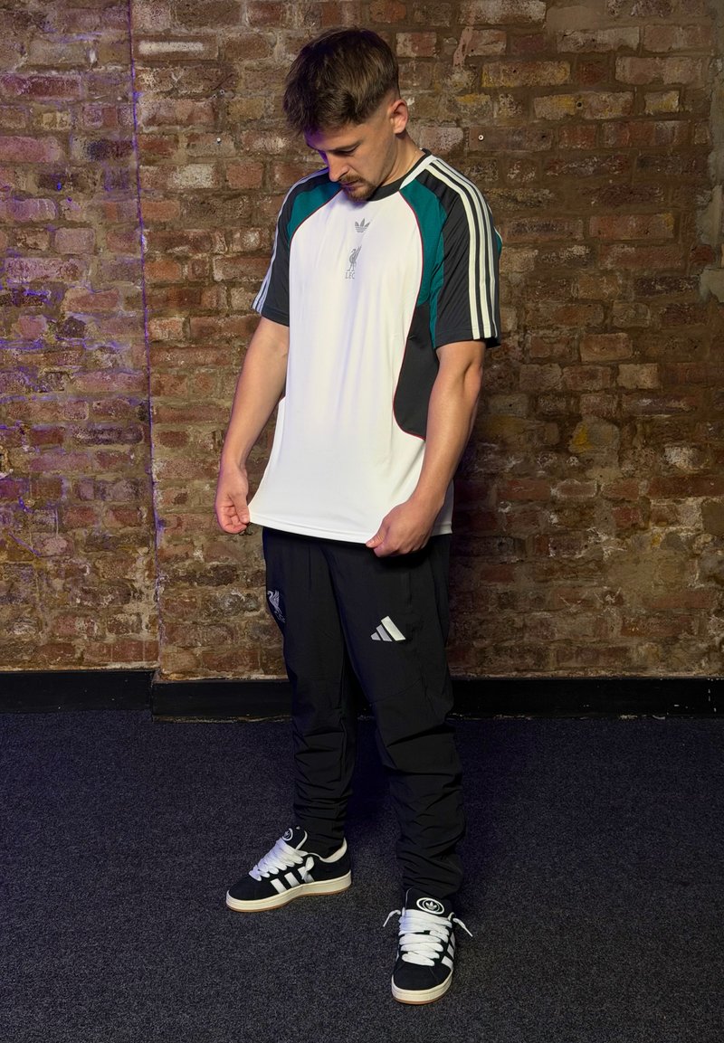 Homme regardant vers le bas, tenant notre de T-shirt de sport blanc et noir, debout devant un mur en briques, portant un pantalon noir et des baskets noires avec des lacets blancs.