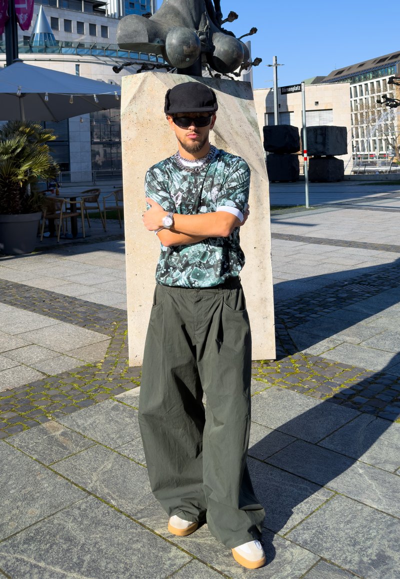 Young man wearing a black cap, sunglasses, patterned shirt, and wide green pants stands with arms crossed near a stone sculpture in an urban plaza.