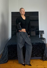 Femme aux cheveux plaqués en arrière portant un large blazer noir, un pantalon gris ample et des talons pointus noirs, debout dans une chambre avec un lit noir et une lampe.