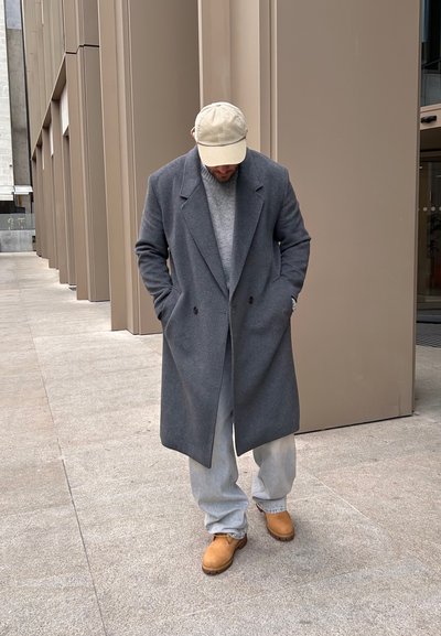Abrigo de lana gris con solapas en pico, llevado sobre un suéter gris. Jeans de corte holgado en azul claro y zuecos color beige. Fondo de piedra en tonos neutros.
