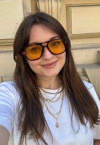 Übergroße Schildpatt-Sonnenbrillen mit gelben Gläsern. Mehrere goldene Ketten und ein weißes T-Shirt. Langes braunes Haar; strukturierter Hintergrund.