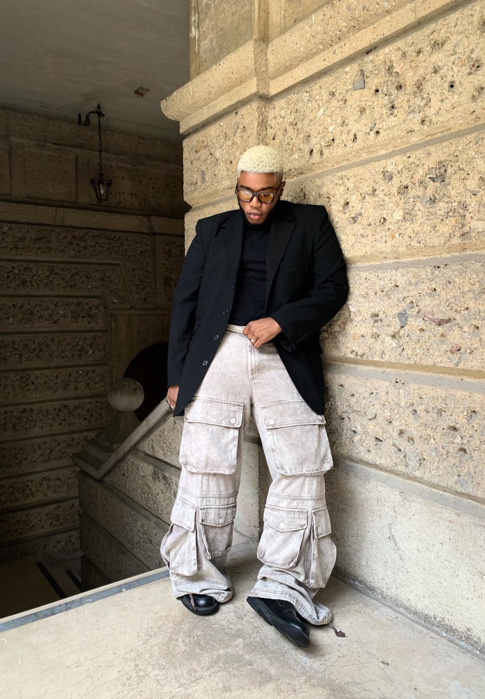 Persona che indossa un blazer nero sopra una camicia nera, pantaloni cargo beige chiaro con tasche e scarpe nere, in piedi contro un muro strutturato.