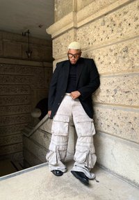 Persona che indossa un blazer nero sopra una camicia nera, pantaloni cargo beige chiaro con tasche e scarpe nere, in piedi contro un muro strutturato.