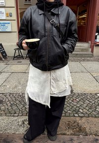 Veste en cuir noire avec une fermeture éclair, jupe blanche à plusieurs couches avec des accents en dentelle, et pantalon noir ample ; tenant une tasse de café noire.