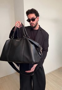 Homme en costume noir et lunettes de soleil tient un grand sac de voyage en cuir noir à l'intérieur, devant un mur blanc uni.