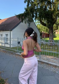 Top halter ajustado em lantejoulas rosas, combinado com calças largas de perna larga rosa claro. Cabelo preso com um prendedor branco, de pé ao ar livre, perto de uma casa.