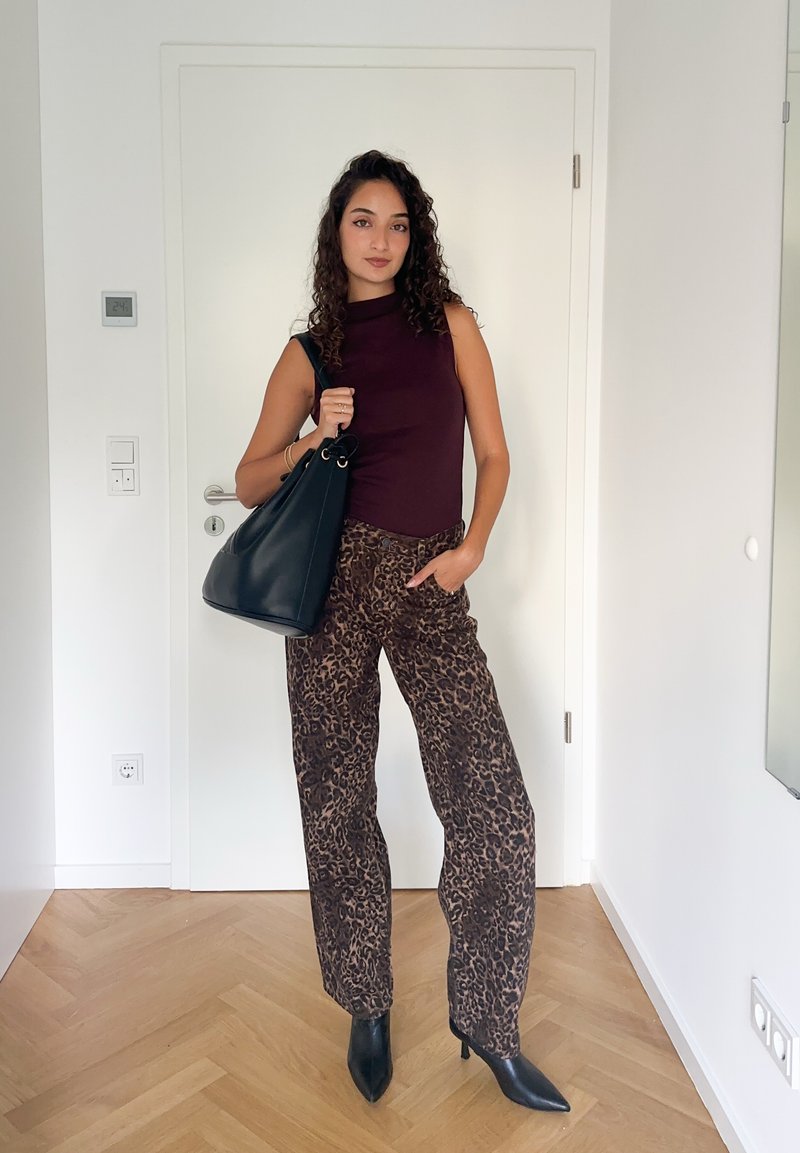 Femme aux cheveux bouclés portant un top sans manches bordeaux, un pantalon à imprimé léopard, des bottes noires à bout pointu et portant un grand sac à main noir à l'intérieur.