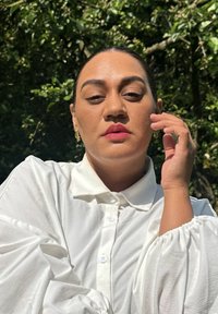 Weißes Langarmhemd mit Knopfkragen und voluminösen Ärmeln. Die Hand des Models ist an das Gesicht gehoben, umrahmt von einem grünen Hintergrund und Sonnenlicht.