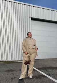 Μπεζ κοστούμι προσαρμοσμένο με πλεκτό σακάκι, συνδυασμένο με μια μικρή τσάντα χρώματος καφέ και μαύρου με μοτίβο. Λευκά παπούτσια ολοκληρώνουν την εμφάνιση σε γκρίζο φόντο.