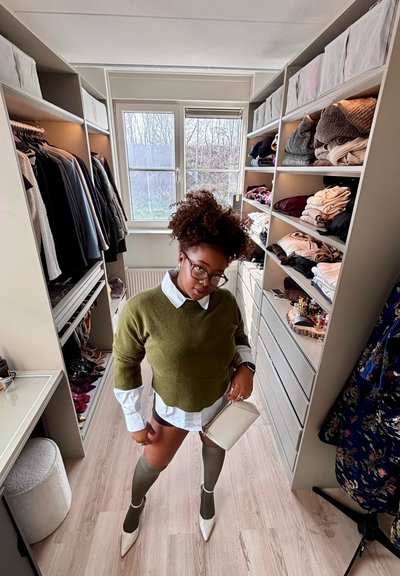 Mujer con suéter verde, camisa blanca y calcetas altas de pie en un vestidor con ropa doblada y prendas colgadas en estantes.