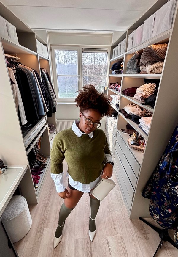 Kobieta w zielonym swetrze, białej koszuli i wysokich skarpetach stoi w garderobie z ułożonymi na półkach złożonymi ubraniami i wiszącymi na wieszakach.