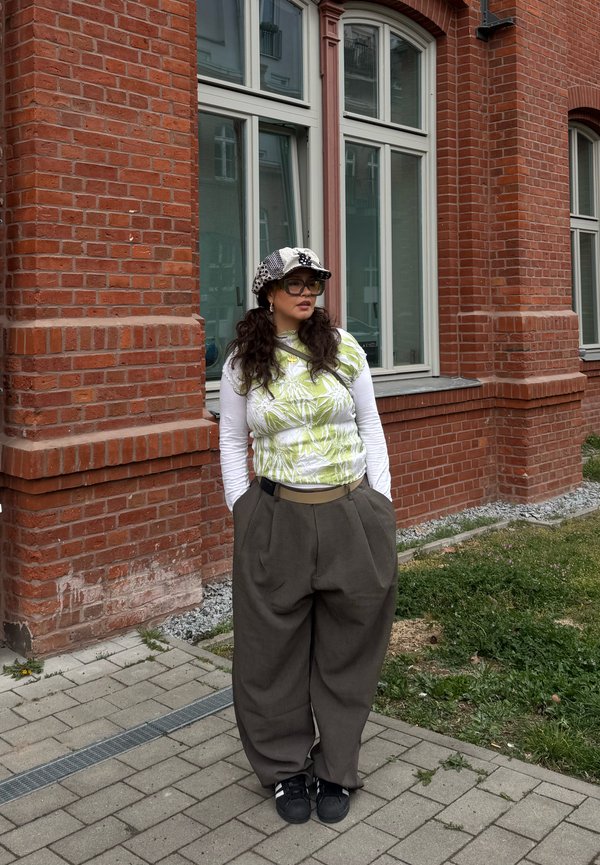 Femme aux longs cheveux foncés portant une casquette à motifs, des lunettes, un haut blanc à manches longues sous un top vert imprimé, un pantalon large gris et des chaussures noires, debout près d'un bâtiment en briques.