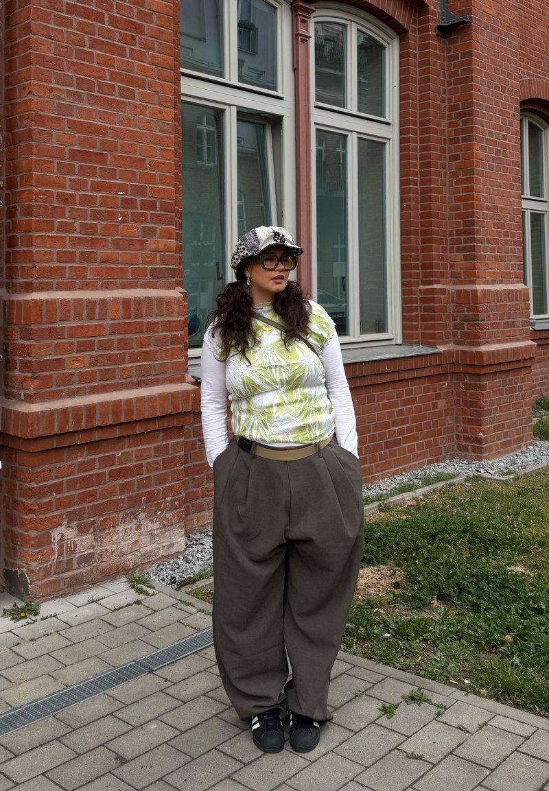 Femme aux longs cheveux foncés portant une casquette à motifs, des lunettes, un haut blanc à manches longues sous un top vert imprimé, un pantalon large gris et des chaussures noires, debout près d'un bâtiment en briques.
