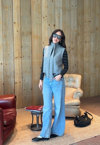 Mujer con gafas de sol oscuras, suéter gris sin mangas con bufanda, camiseta de manga larga a rayas, pantalones vaqueros anchos azules, de pie en una habitación de madera con sillas y bolso.