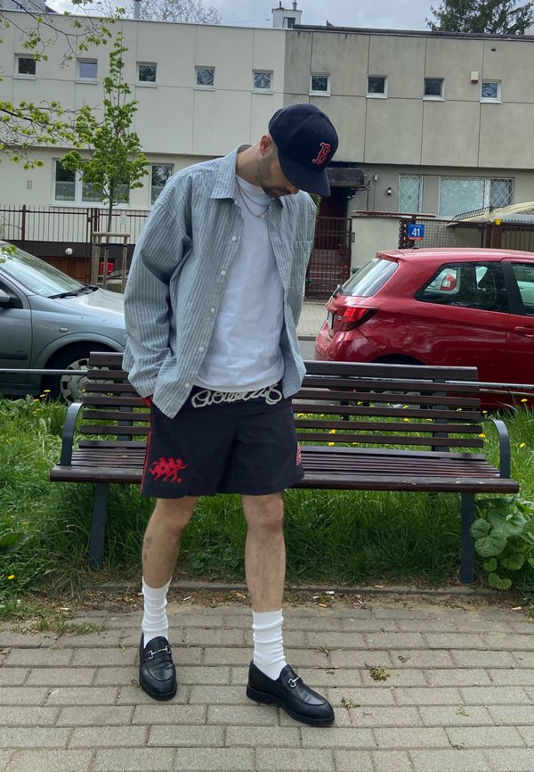 Man met een zwarte pet, grijs overhemd over een wit T-shirt, zwarte korte broek, witte sokken en zwarte loafers, staand op een geplaveid pad nabij een parkbank.