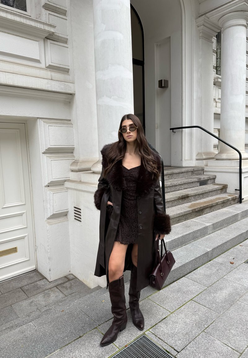 Abrigo trench de piel marrón con cuello de piel, vestido marrón texturizado y botas hasta la rodilla. Sosteniendo un bolso burdeos oscuro. Escaleras de piedra en el fondo.