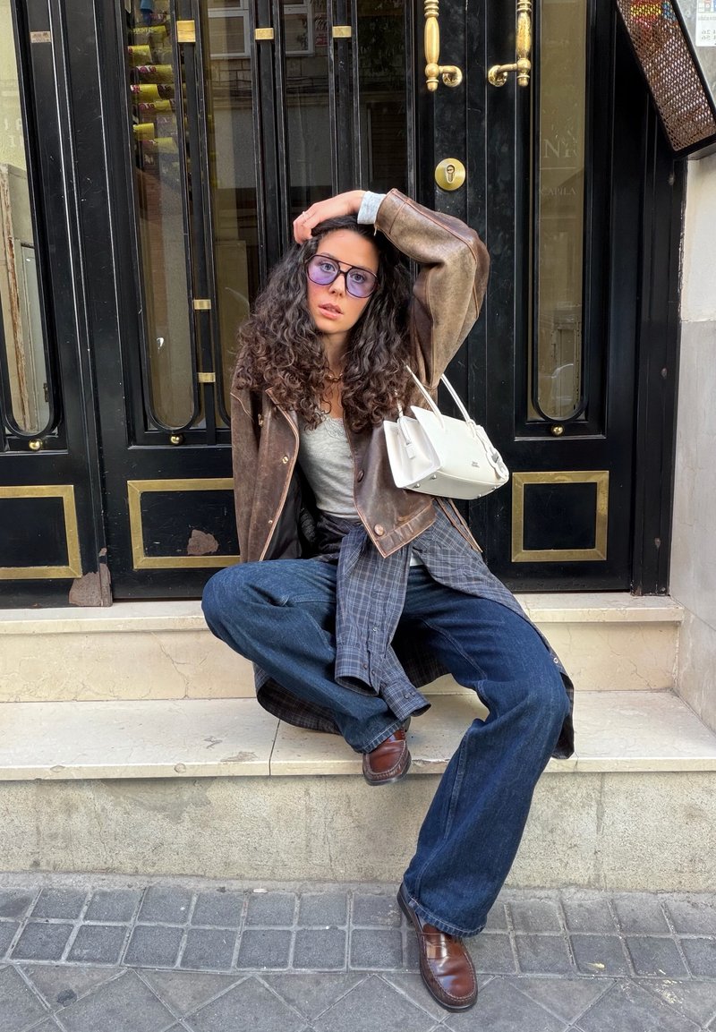Donna con capelli ricci che indossa occhiali colorati di viola, una giacca di pelle marrone, una maglietta grigia e jeans larghi, seduta su gradini di pietra davanti a porte doppie nere.