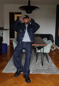 Donkerblauwe spijkerjack met contrasterende stiksels, grijze sweatshirt, losse blauwe spijkerbroek en zwarte schoenen. Binnenomgeving met meubels.