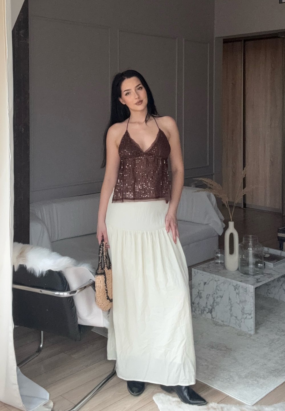 Donna in top a fascia marrone di paillettes e gonna bianca lunga, con una borsa intrecciata in mano, che si trova in un soggiorno moderno con un tavolino da caffè in marmo.
