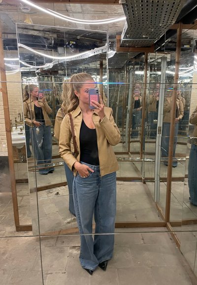 Chaqueta de cuero marrón, blusa negra y vaqueros de pierna ancha azules. Zapatos negros de puntera. Reflejos en un espacio lleno de espejos. Iluminación brillante en el techo.