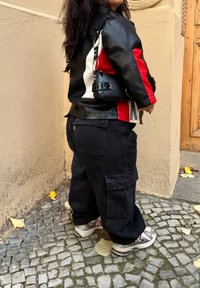 Giacca di pelle nera con accenti rossi e bianchi, abbinata a pantaloni cargo neri larghi e scarpe sportive argentate su una superficie di ciottoli texturizzati.