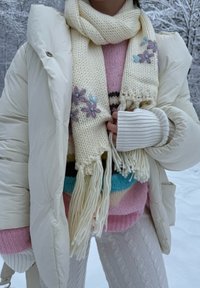 Cachecol de lã creme com bordados florais, combinado com um suéter rosa claro e uma jaqueta acolchoada branca. Look texturizado e em camadas.