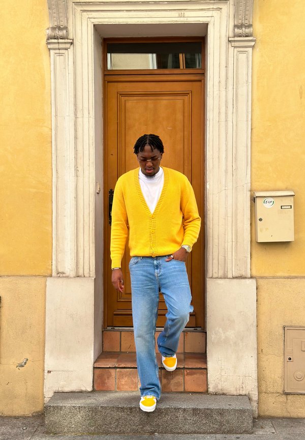 Man wearing yellow sweater, white shirt, blue jeans, and yellow shoes stepping down from a doorway with framed stone arch and beige walls.