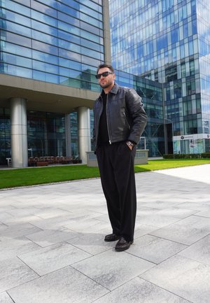 Chaqueta de cuero negra, camisa negra, pantalones holgados negros y zapatos marrones; de pie sobre un pavimento de piedra con un edificio de cristal de fondo.