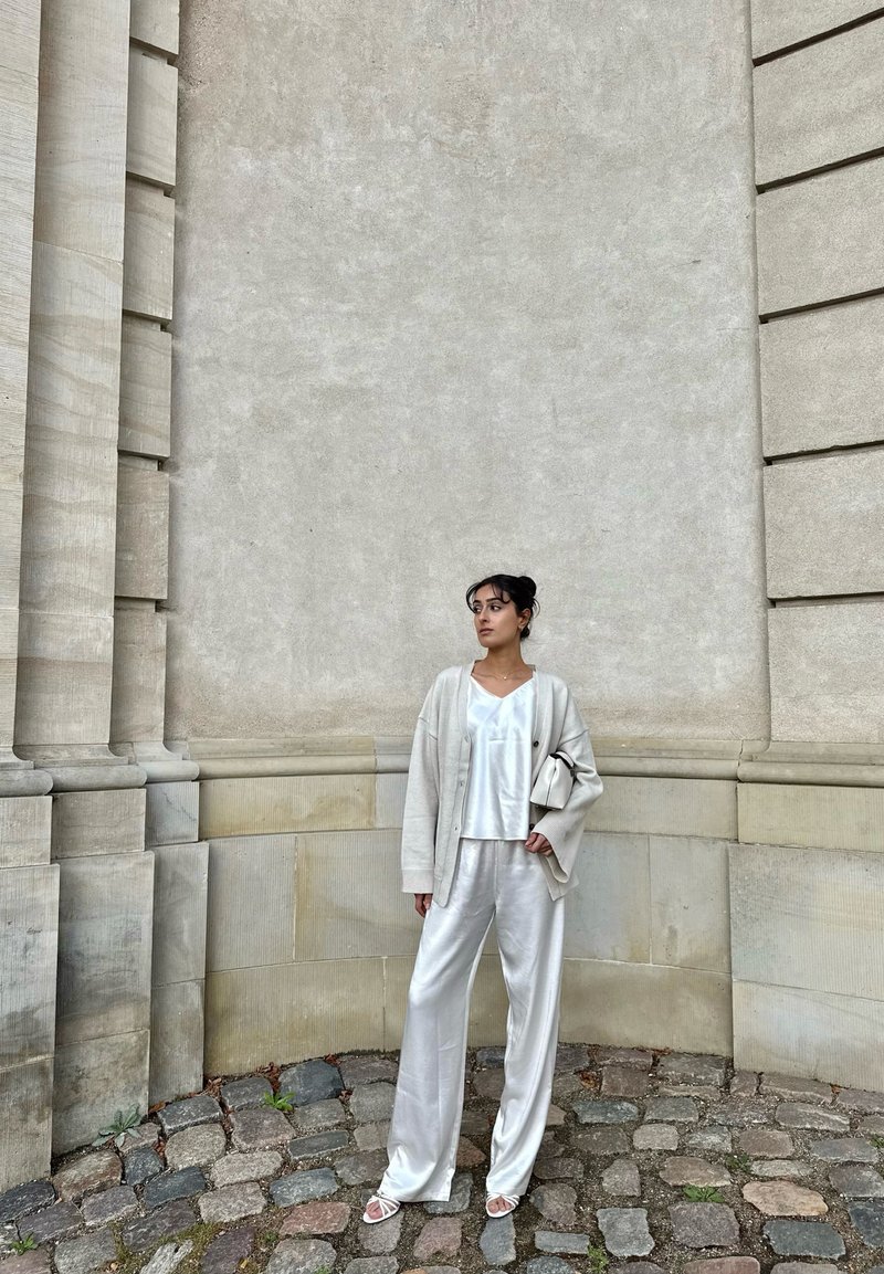 Woman in a white satin outfit and beige cardigan stands on a cobblestone street with a large stone wall in the background.