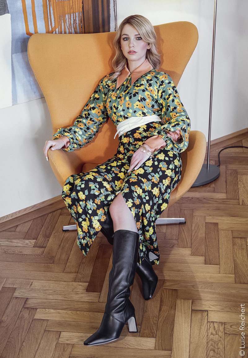 Floral patterned dress in green, yellow, and black; orange collar; white belt. Black knee-high boots. Sits on an orange sculptural chair.