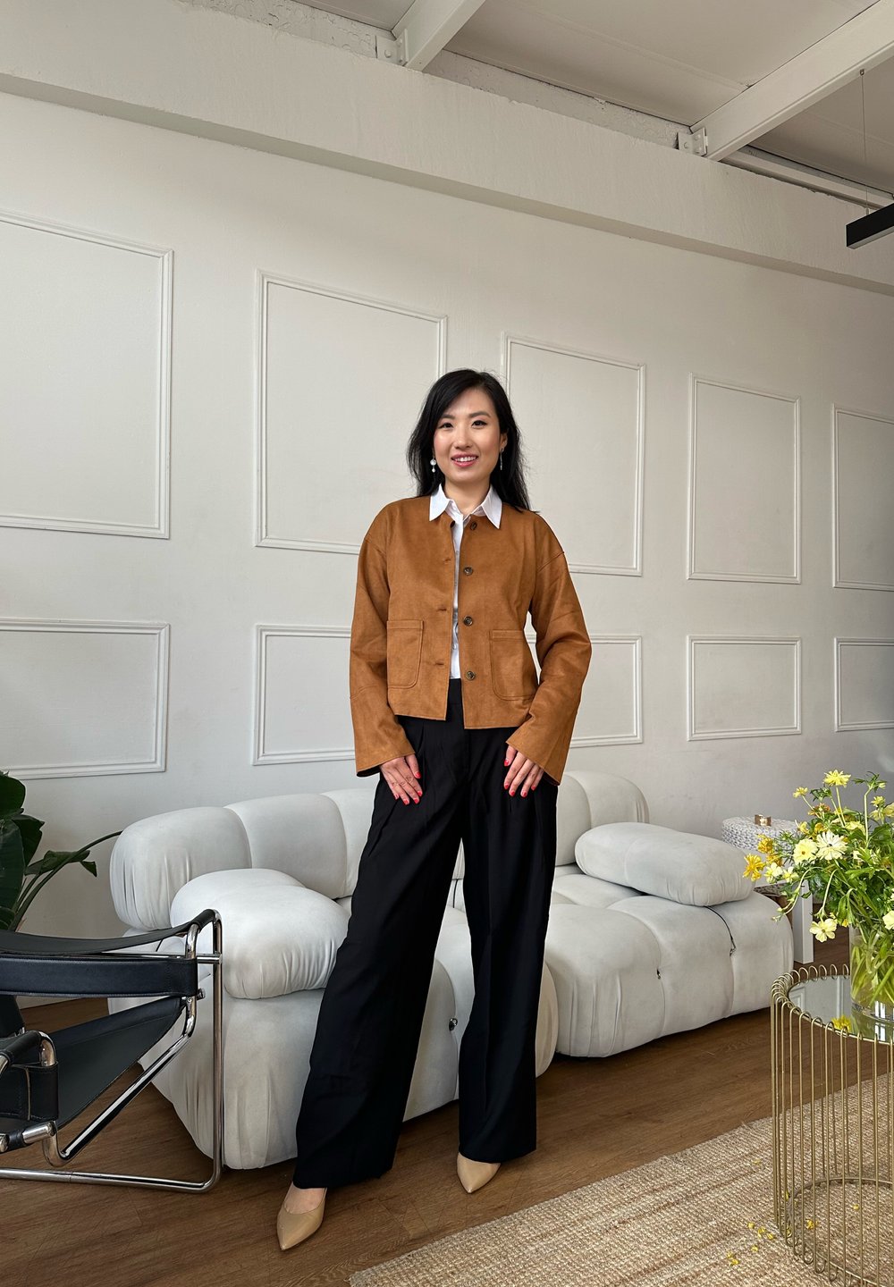 Veste en daim marron avec col rond et poches avant, associée à un pantalon noir à jambes larges et des escarpins beiges à bout pointu dans un intérieur moderne.