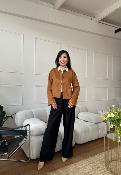 Chaqueta de ante marrón con cuello redondo y bolsillos delanteros, combinada con pantalones anchos negros y tacones beige de punta puntiaguda en un interior moderno.