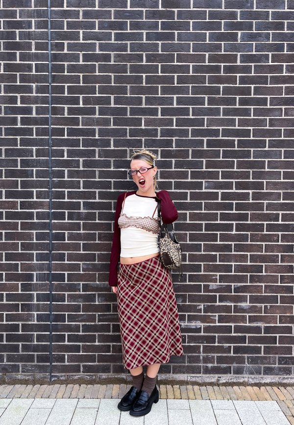Une femme portant des lunettes et une jupe à carreaux pose la bouche ouverte devant un mur de briques sombre, avec un sac à bandoulière à motifs.