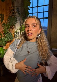 Sølvstjernehalskjede, grå kortermet skjorte, hvit langermet overlay, intrikate ringer, krøllete hår, innendørs setting med planter og vindu.