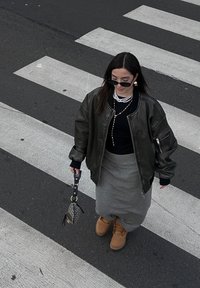 Πράσινο bomber μπουφάν, μαύρη μπλούζα, γκρίζα φούστα και ταμπα μπότες. Αξεσουάρ περιλαμβάνουν ένα κολιέ με πέρλες και μία μοτίβο τσάντα. Φόντο από διάβαση πεζών.