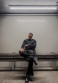 Chemise noire à manches longues avec un col boutonné, pantalon large noir, et chaussures noires. Assis sur un banc gris devant un fond uni.